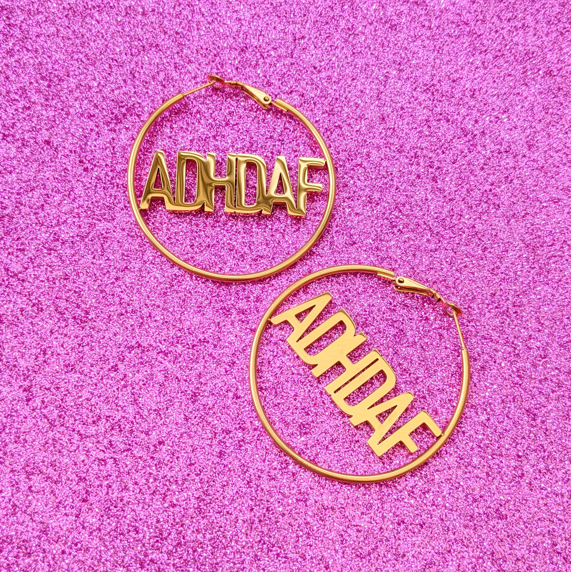 Gold Hoop Earrings with letters ADHDAF across middle on a pink glittery background