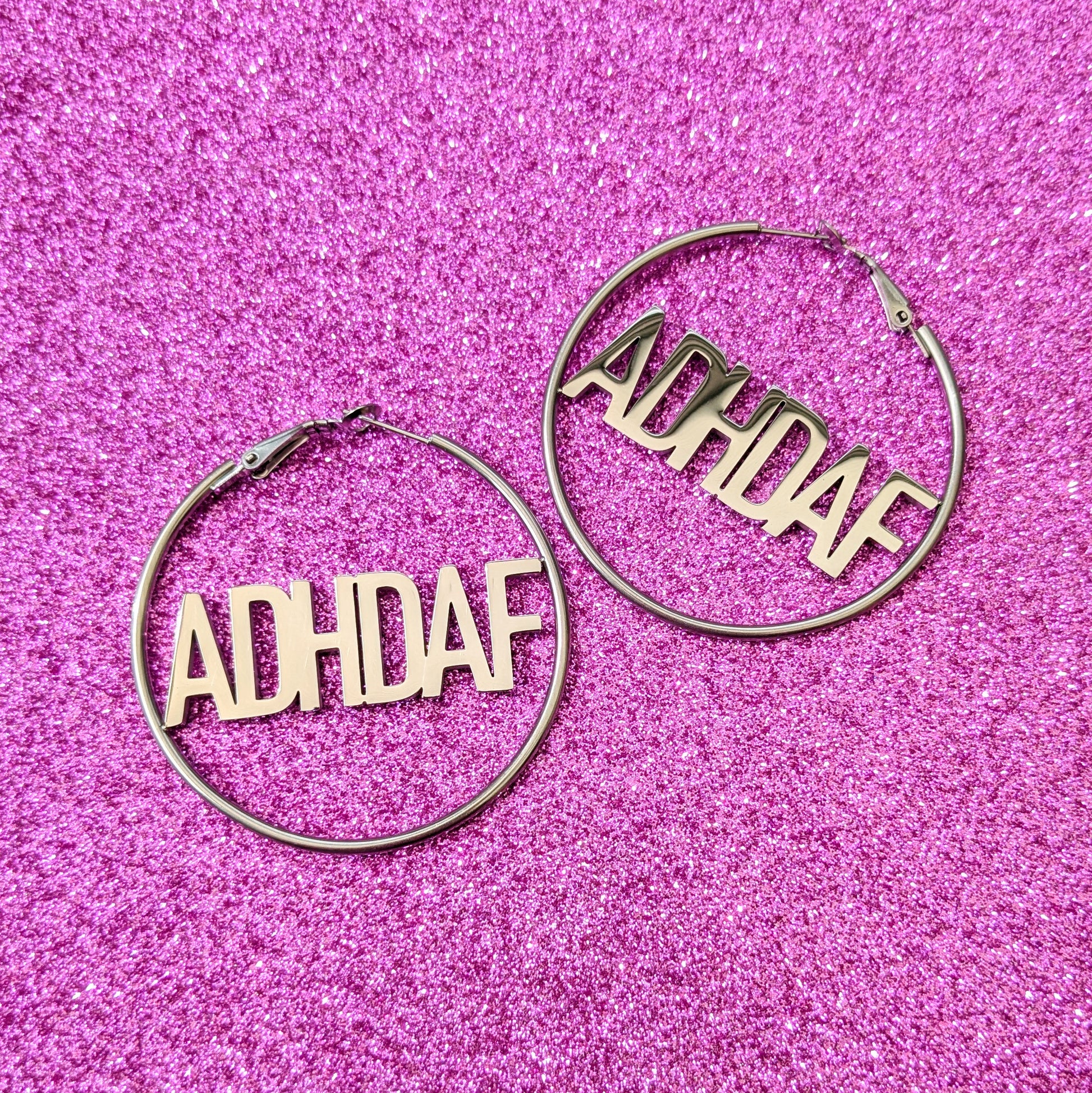 Silver Hoop Earrings with letters ADHDAF across middle on a pink glittery background