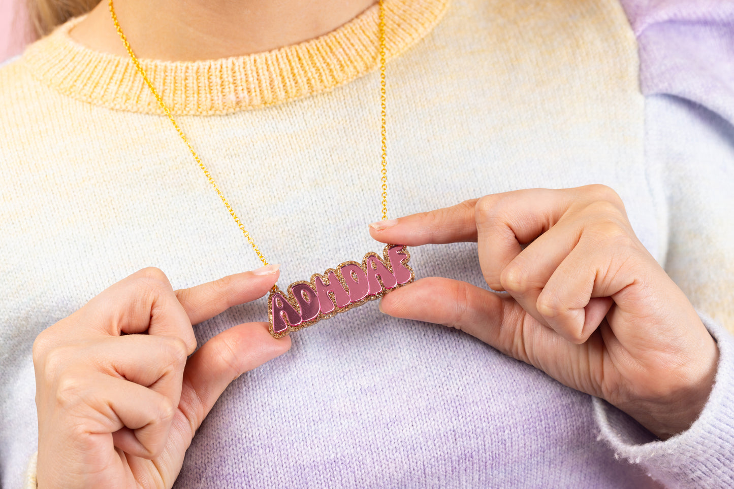 Model wears pink ADHDAF Necklace on a gold chain, she holds the necklace on the corners of the letters