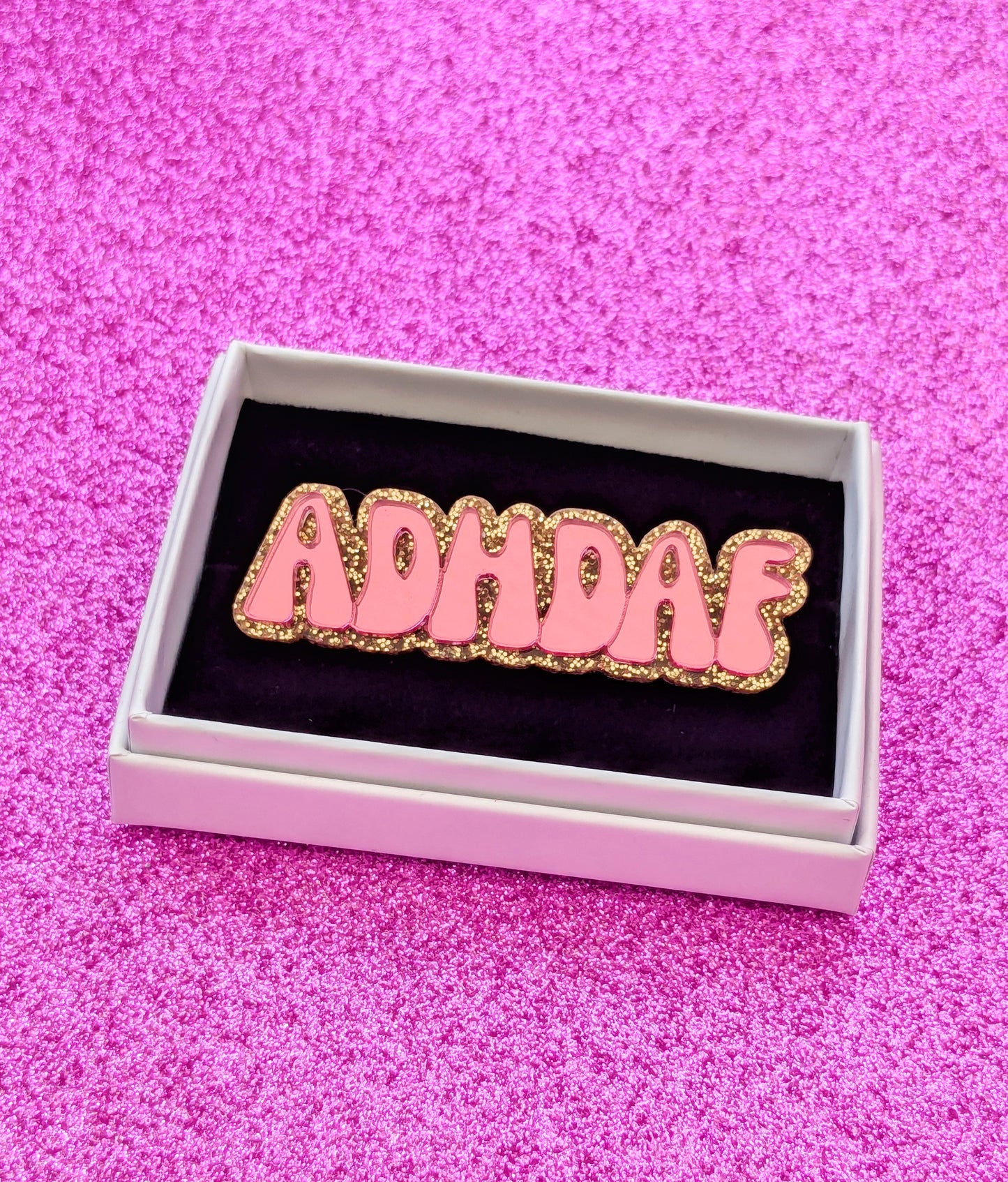 Pink acrylic ADHDAF pin framed in a white box on a glittery pink background 