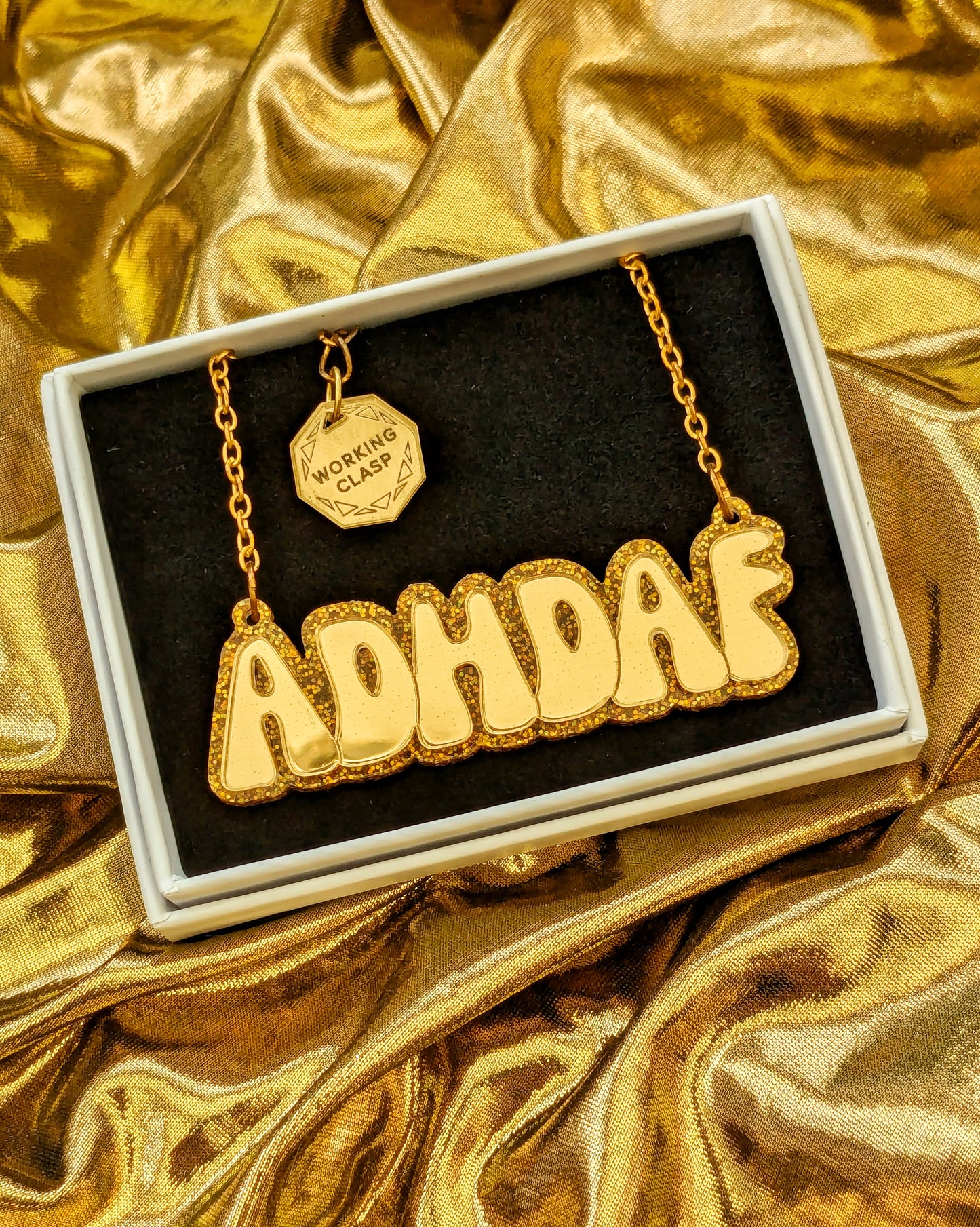 ADHDAF acrylic necklace in gold on a gold chain boxed on a gold fabric background