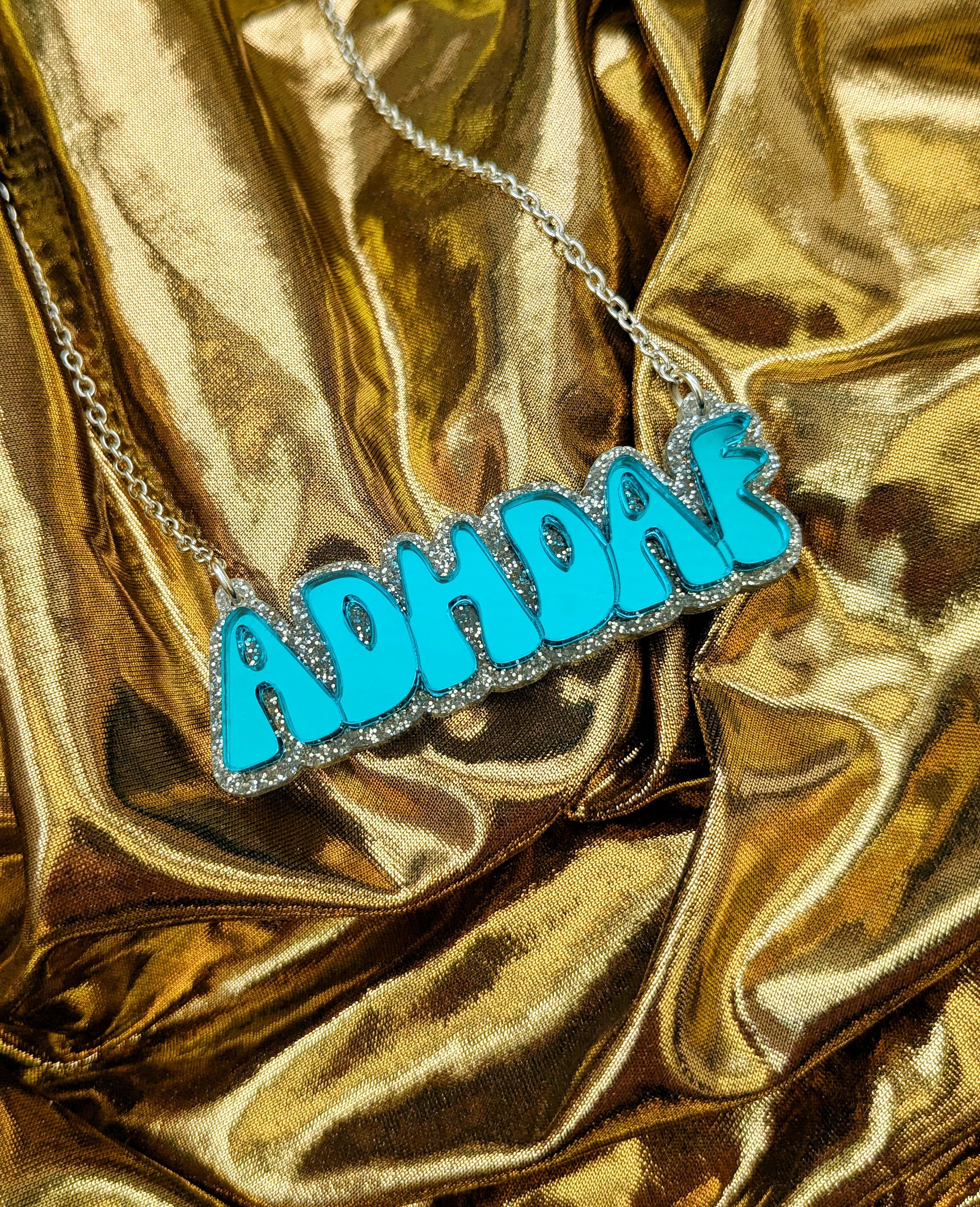 ADHDAF acrylic necklace in turquoise on a silver chain boxed on a gold fabric background