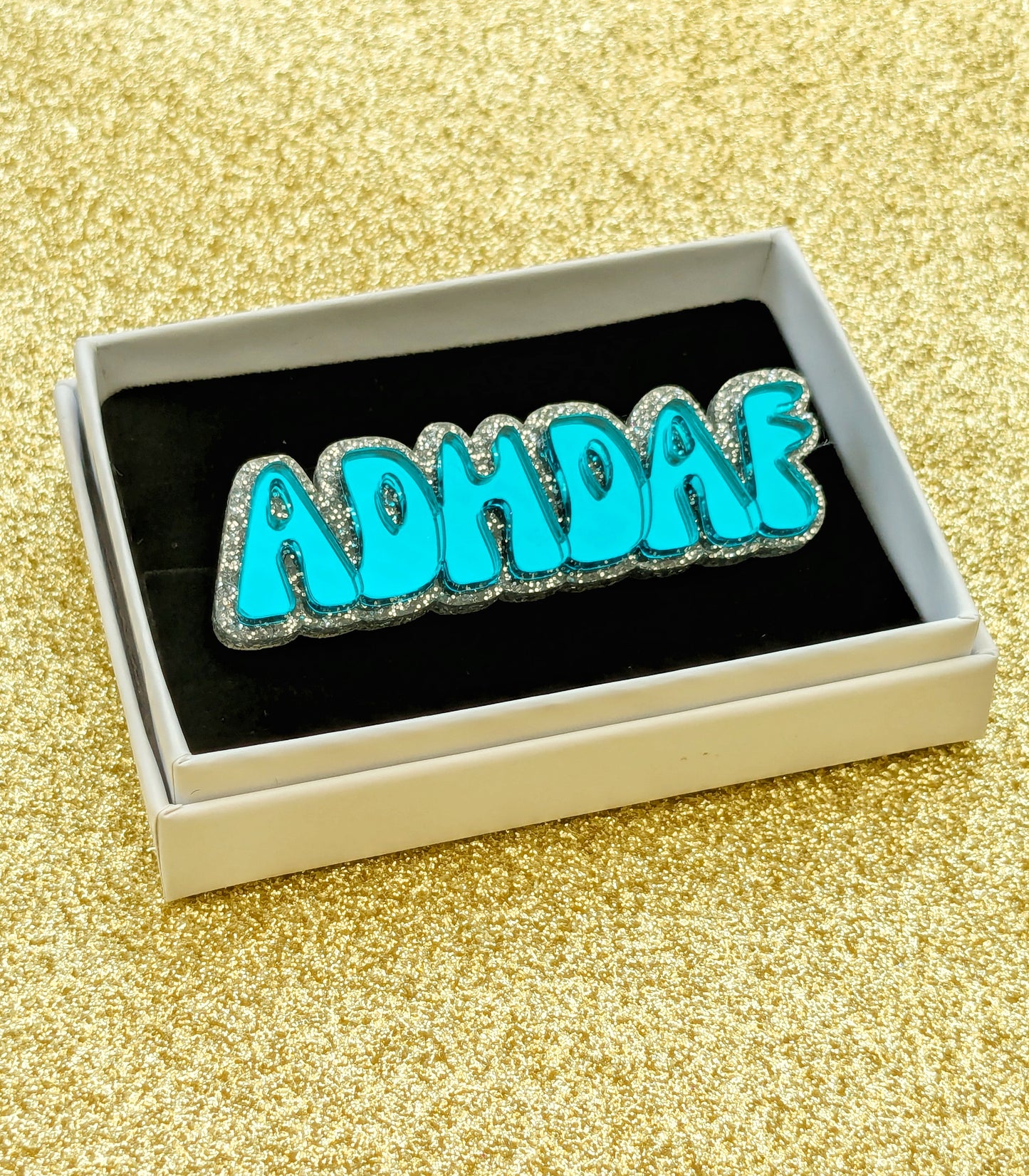 Turquoise acrylic ADHDAF pin in a white box on a glittery gold background