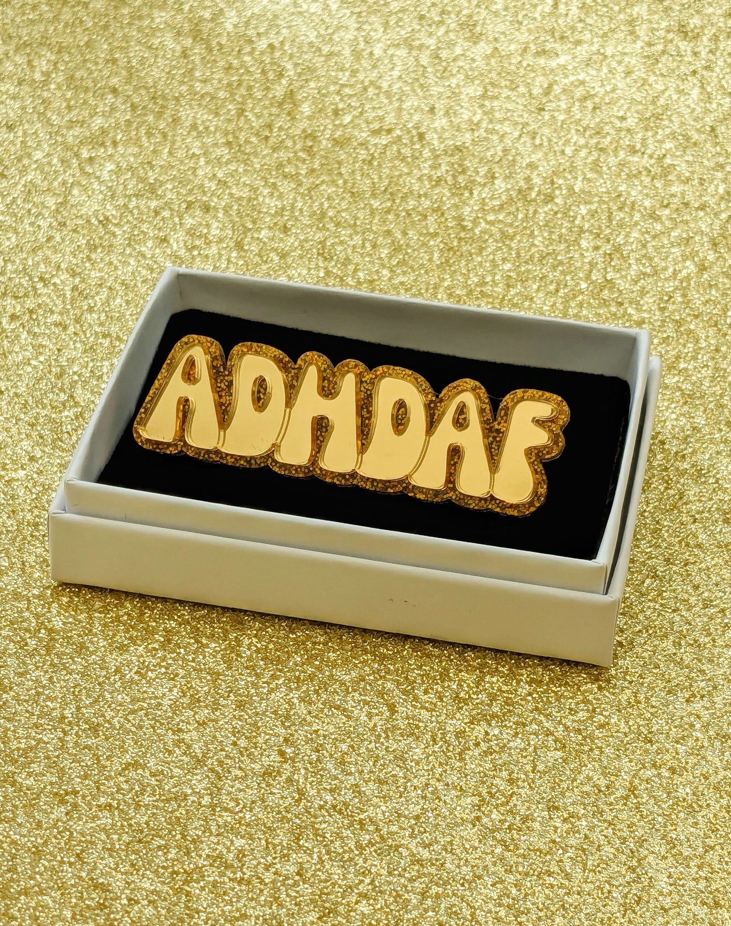 Gold acrylic ADHDAF pin in a white box on a glittery gold background