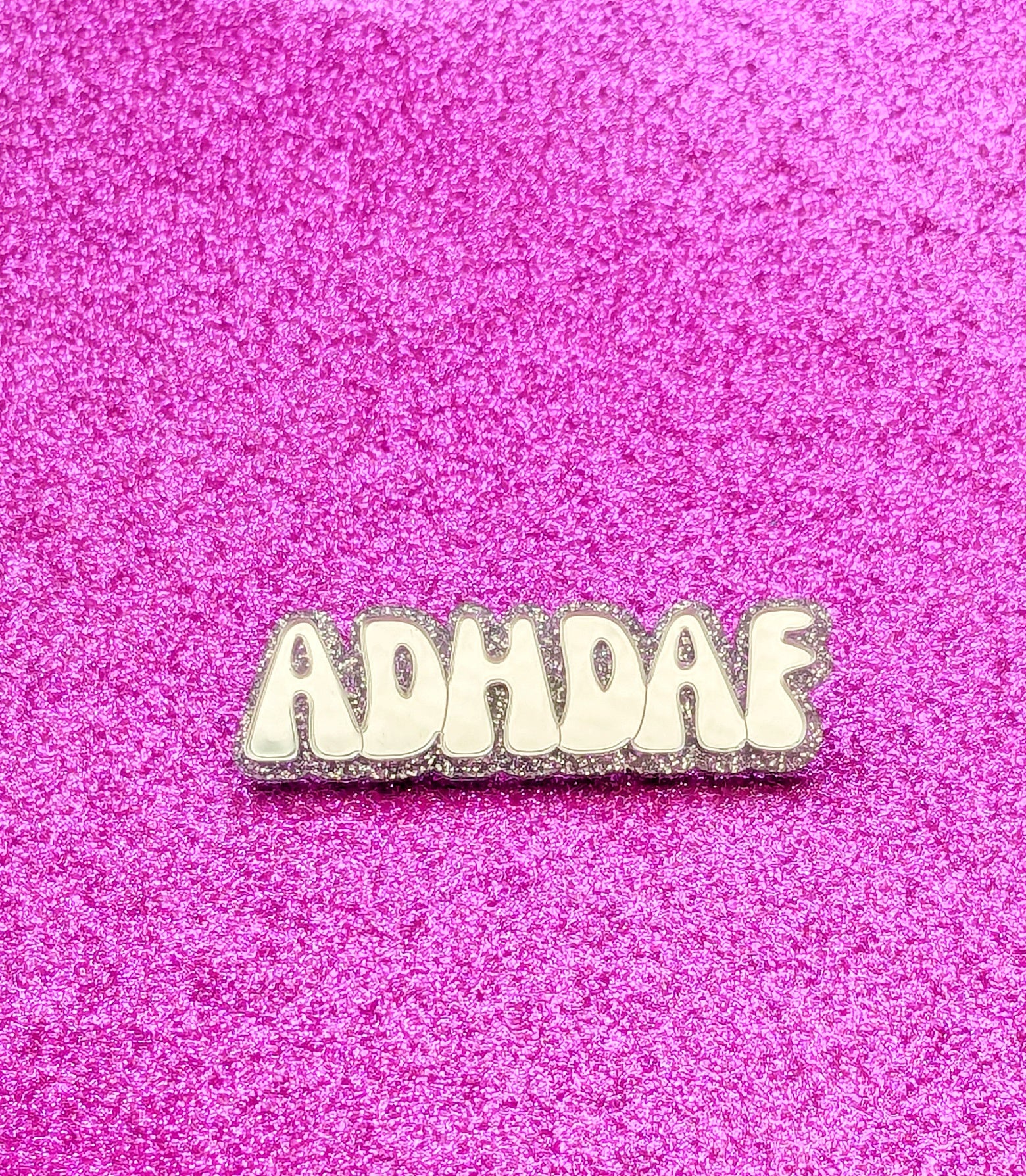 Silver acrylic ADHDAF pin on a glittery pink background