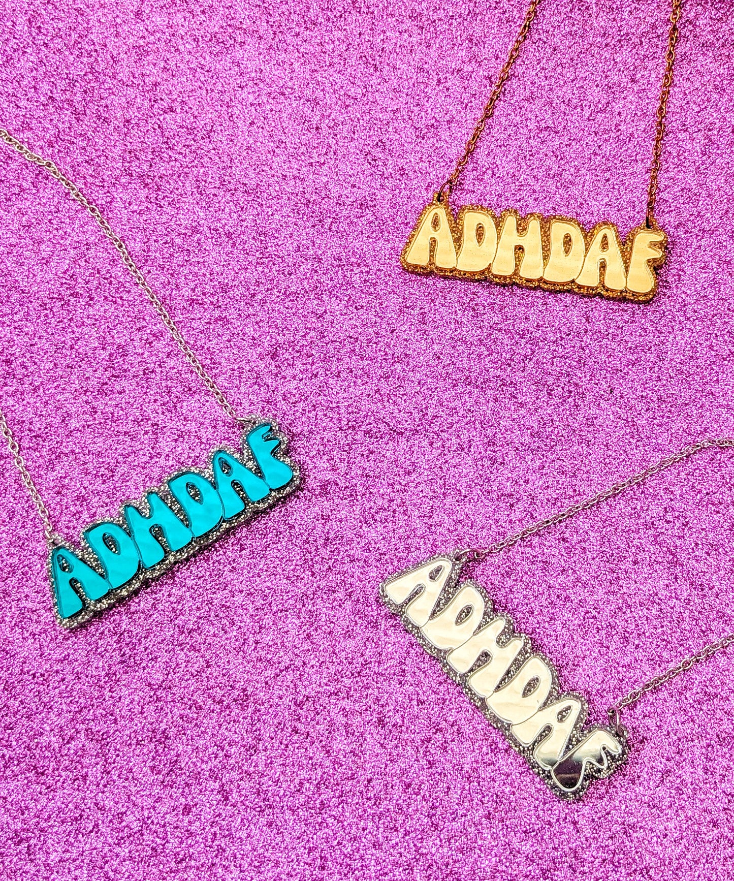 Display of 3 ADHDAF necklaces in Gold, Turquoise and Silver on a sparkly pink background