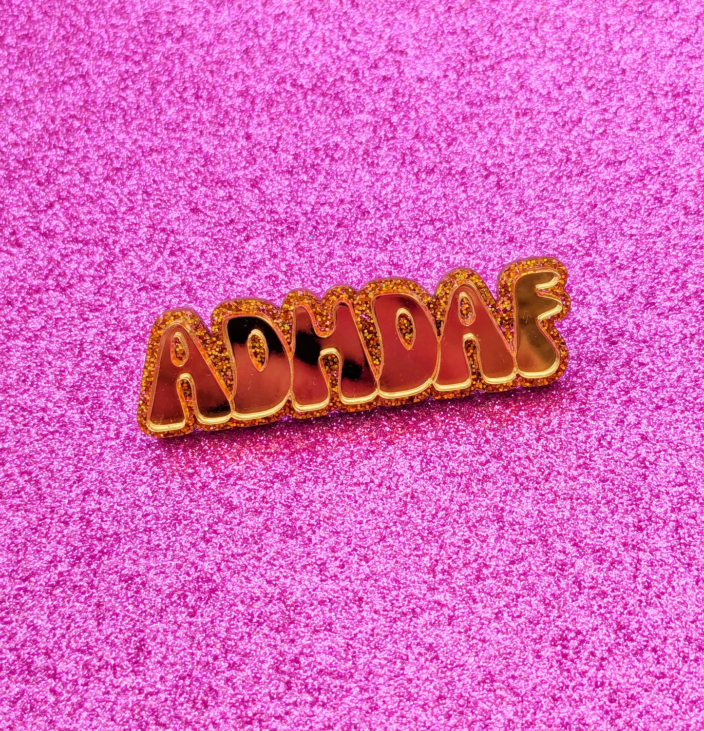 Gold acrylic ADHDAF pin on a glittery pink background