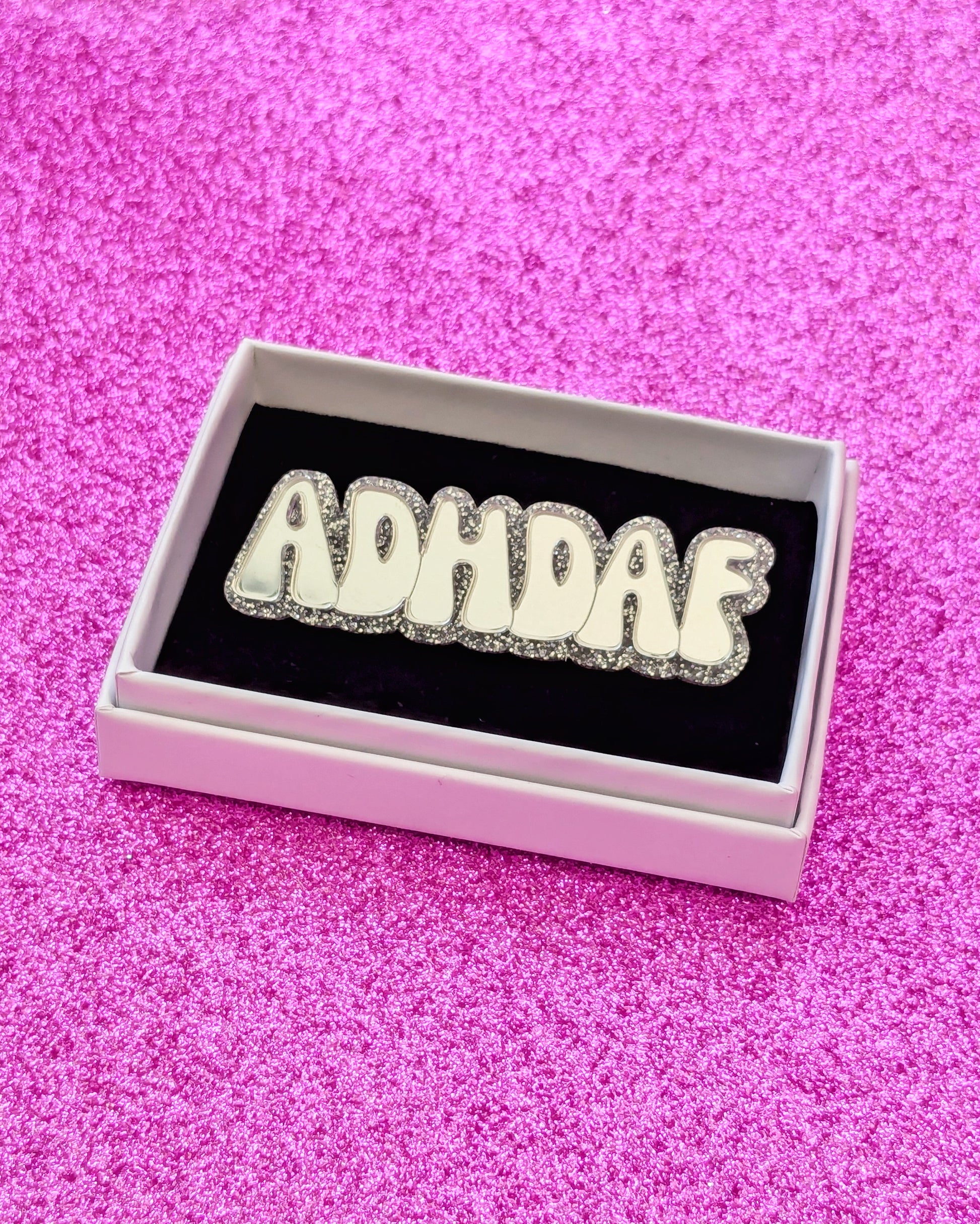 Silver acrylic ADHDAF pin in a white box on a glittery pink background