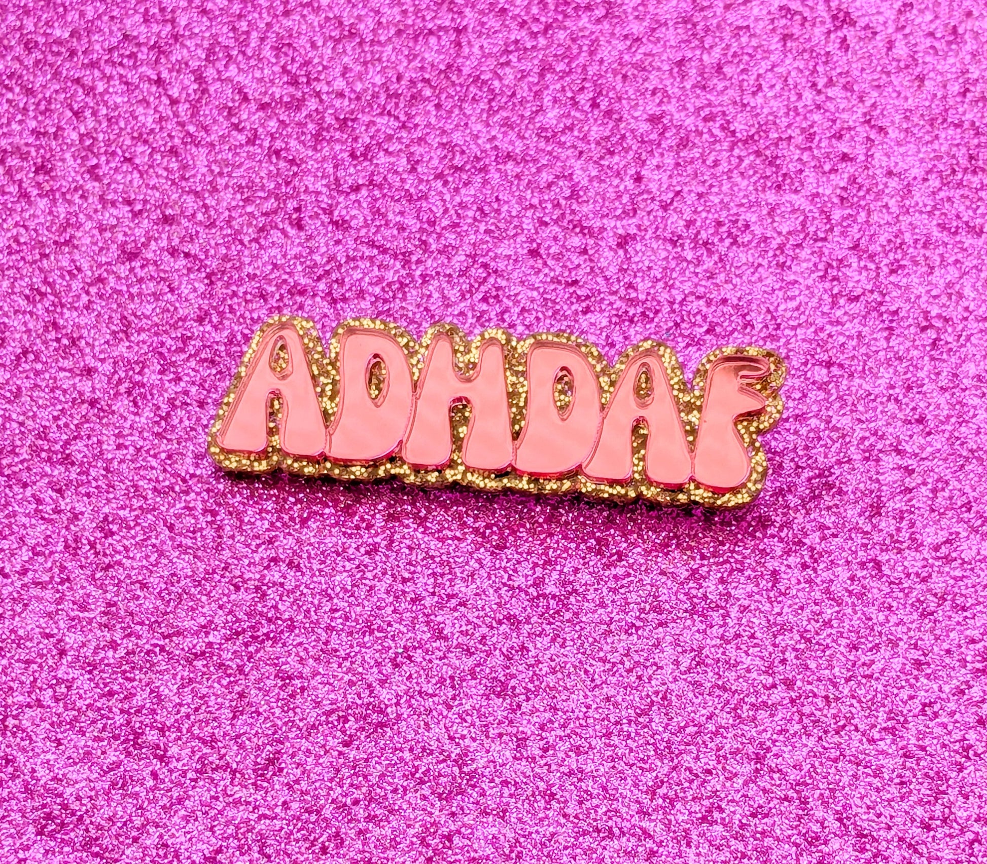 Pink acrylic ADHDAF pin on a glittery pink background